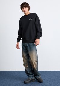 Black sweatshirt with logo, loose-fit, paired with faded blue baggy jeans featuring distressing. Model wears chunky black clogs.