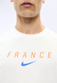 Κοντινό πλάνο ενός ατόμου που φορά λευκό πουκάμισο με τη λέξη «FRANCE» σε πορτοκαλί γράμματα και το μπλε λογότυπο Nike κάτω από το κείμενο.