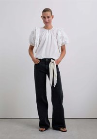 Weiße Bluse mit Rüschensäumen, kombiniert mit schwarzen Schlagjeans. Das Outfit enthält einen cremefarbenen Stoff, der um die Taille gebunden ist. Glatte Texturen.