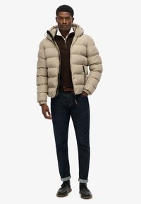 Beige pufferjack met een capuchon, ritssluiting en gewatteerd ontwerp. Draag het over een bruine trui en combineer het met donkere denim en zwarte schoenen.