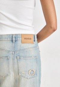 Jeanși din denim albastru deschis au o etichetă din piele inscripționată "HUGO" la spatele taliei, cu accente uzate și un logo circular brodat pe un buzunar.