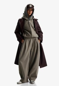 Hanorac gri supradimensionat și pantaloni de trening asortați cu un trench din piele maro și încălțăminte argintie. O șapcă cu logo pe față completează ținuta.
