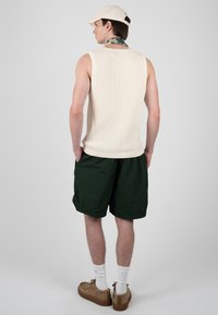 Jeune homme portant un débardeur tricoté blanc, un short vert foncé, une casquette beige, un foulard à motifs autour du cou, des chaussettes blanches et des chaussures marron, vu de dos.