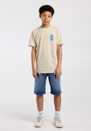 Jongen staand, draagt een beige T-shirt met print, denim shorts, witte sneakers en witte sokken met blauwe strepen tegen een effen achtergrond.