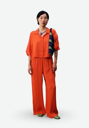 Vrouw draagt een loszittend oranje overhemd met knopen en wijde broek, groene geweven schoenen, gestreepte schoudertas en een kralenketting met blauwe hartvormige hanger.