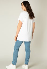 Witte, T-shirt met korte mouwen en een ontspannen pasvorm, gecombineerd met lichtblauwe spijkerbroek en witte sneaker. Achteraanzicht toont een ronde halslijn.