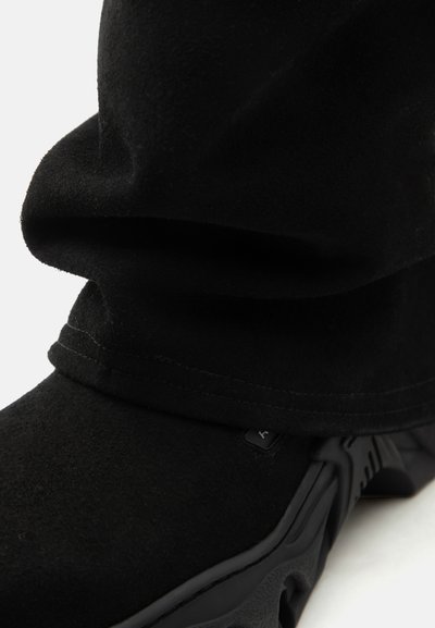 Μαύρο μπότες από σουέντ με διπλωμένη μανσέτα και παχύ, υφασμένο καουτσούκ σόλα, ορατή σε λευκό φόντο.