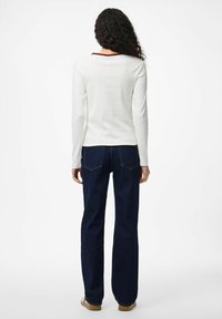 Kvinna med långt lockigt hår som bär en vit långärmad topp, mörkblå jeans och bruna skor, stående mot en enfärgad vit bakgrund.