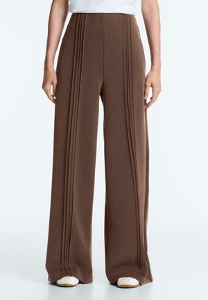 Femme portant un pantalon marron taille haute à jambes larges avec des plis verticaux, associé à des baskets blanches et un haut blanc.