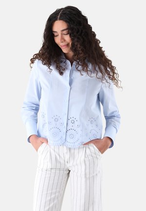 Frau mit langen lockigen Haaren, die ein hellblaues Hemd mit Knöpfen und ausgeschnittenen Blumenmustern sowie weiße Hosen mit dünnen vertikalen Streifen trägt.