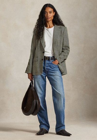 Vrouw met lang krullend haar, een visgraatblazer, wit T-shirt, blauwe spijkerbroek, zwarte laarzen, die een grote bruine handtas vasthoudt in een neutrale studio-omgeving.