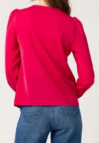 Fitted long-sleeve top in bright pink fabric with a smooth texture and puffed shoulders. Paired with blue denim jeans featuring back pockets.