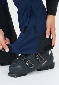 Svarta skidor med justerbara spännen, i kombination med marinblå vattenavvisande byxor med svarta detaljer och kardborrestängning vid fållarna.