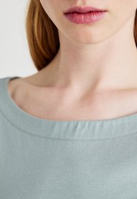 Top din bumbac moale, de culoare albastru deschis, cu un decolteu larg și o textură subtilă. Materialul neted este ușor plisat la decolteu pentru un plus de detaliu.