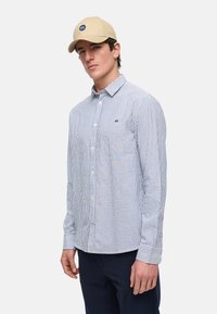 Gestreepte overhemd in blauw en wit, lange mouwen, met een getailleerde pasvorm. Beige pet met logo-detail, tegen een effen achtergrond.
