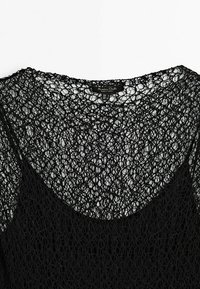 Haut en tricot noir présentant une texture ouverte et semblable à de la dentelle avec un col large. Le design est léger et aéré, idéal pour le superposition.