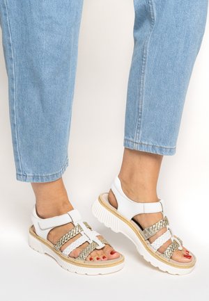Füße in weißen und goldenen Sandalen mit geflochtenen Riemen und dicken weißen Sohlen, kombiniert mit hellblauen Jeans, die an den Knöcheln gekürzt sind.