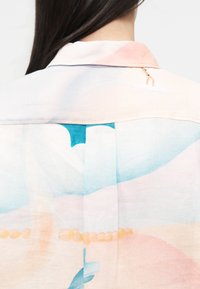 Οπίσθια όψη ενός ατόμου που φοράει πουκάμισο σε παστέλ χρώματα με αφηρημένα σχήματα και μικρό σχέδιο φιγούρας κοντά στον γιακά.