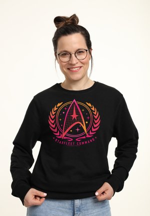 Lächelnde Frau mit Brille und Dutt, die einen schwarzen Starfleet Command Sweatshirt trägt, steht vor einem schlichten hellen Hintergrund.