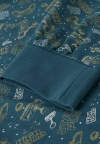 Tissu bleu sarcelle avec poignet côtelé présentant des icônes, boutons et phrases de jeux rétro en or et blanc, tels que "START PLAY" et "HIGH SCORES".