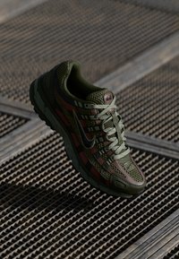 Olive green sneaker with mesh material, patterned overlays, and black accents. Features a lace system and a textured outsole.