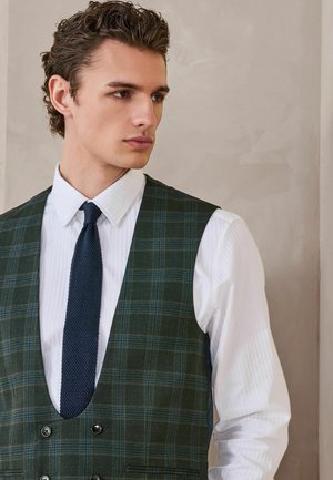 Young man with curly hair wearing a white shirt, navy tie, and green plaid vest, looking to the side against a light wall background.