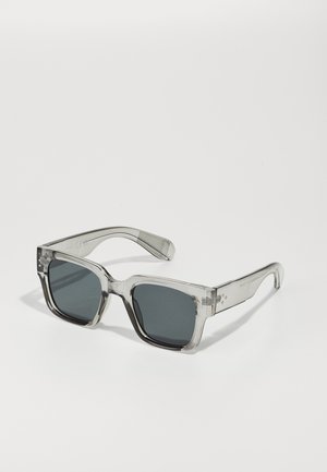 Clear, oversized sunglasses with dark lenses, featuring a chunky rectangular frame and textured temple accents. Made of transparent plastic.