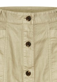 Jupe beige à boutons, avec une taille, deux poches avant et quatre boutons marron. Le tissu présente un aspect lisse et texturé.