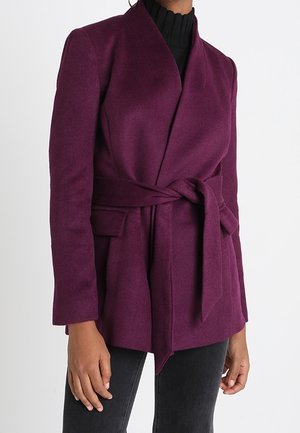 Femme portant un manteau en laine violet profond ceinturé sur un col roulé noir et un pantalon noir, montrée du cou aux hanches.