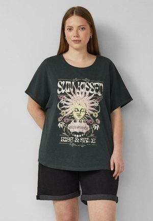 Jonge vrouw met lang haar die hoepeloorbellen draagt, een donker oversized T-shirt met zon- en bloemmotief en tekst, en opgerolde zwarte shorts.