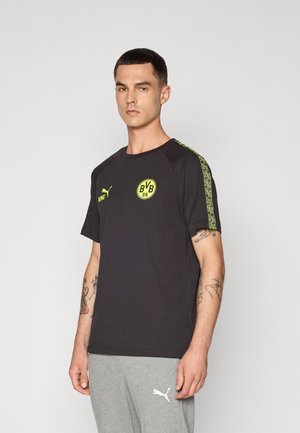 Sort kortærmet t-shirt med gult logo og detaljer. Har mønstret grøn kant rundt om skuldrene. Stoffet ser glat ud; afslappet pasform.