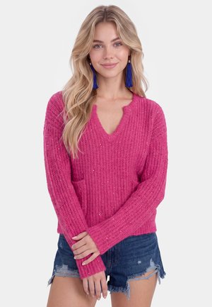 Blond vrouw met een roze gebreide trui met lange mouwen en blauwe spijkershort, glimlachend met handen voor zich geklemd, op een witte achtergrond.