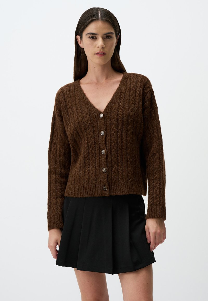 Bruine kabelgebreide cardigan met een V-hals en zes knopen aan de voorkant; ribgebreide manchetten en zoom. Draag een zwarte geplooide rok eronder.