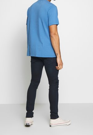 Mann, der von hinten steht, trägt ein hellblaues T-Shirt, dunkle Skinny-Jeans, weiße High-Top-Sneaker und ein Unendlichkeitstattoo an seinem rechten Handgelenk.