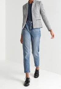 Svetle šedý blazer s kulatým výstřihem, spárovaný s modrými džíny s vysokým pasem a černými šněrovacími botami. Jednoduchý a strukturovaný design.