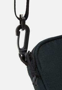 Czarna materiałowa saszetka o gładkiej fakturze, z czarnym plastikowym karabinkiem i plecionym detalem sznura. Widoczny zamek błyskawiczny.