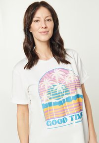 Hvid t-shirt med korte ærmer, der har et farverigt grafisk print med palmer, solnedgangsstriber og teksten "GOOD TIMES" nederst.