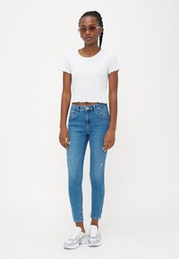 Camiseta corta blanca con textura acanalada y bordes en forma de ondas, combinada con jeans ajustados azul claro con ligeros desgastes, y zapatillas grises.