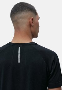 Juodas sportinis marškinėlis su tekstūruotu audiniu, raglaninės rankovės ir vertikali atšvaitinė detalė ant nugaros matomumui.
