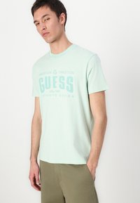 Camiseta de algodón verde claro con cuello redondo, mangas cortas y texto impreso "GUESS" en un verde ligeramente más oscuro. Ajuste casual, sin patrones visibles.