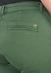Pantalon vert en tissu doux, doté d'une poche arrière avec des coutures et d'un petit logo doré accrocheur. Texture lisse avec une coupe ajustée.