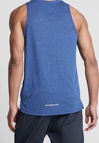 Widok z tyłu osoby w niebieskiej bezrękawnikowej koszulce sportowej i czarnych szortach, stojącej na gładkim jasnym tle.