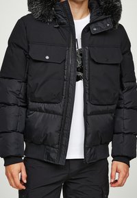 Schwarze Daunenjacke mit abnehmbarem, mit Fell gefüttertem Kapuzenkragen, zwei Fronttaschen, Druckknöpfen und gerippten Bündchen. Strukturierter, isolierender Stoff.