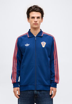 Giovane uomo con giacca blu con zip, con strisce rosse sulle maniche e stemma del calcio croato sul petto, in piedi su sfondo bianco.