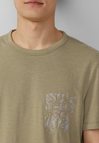 Koszulka w oliwkowym kolorze z teksturowanym motywem liści na lewej kieszeni; okrągły dekolt i krótkie rękawy.