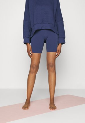 Osoba stoi boso na różowej macie do jogi, ubrana w granatowe spodenki kolarskie i luźną granatową bluzkę z długim rękawem, na białym tle.