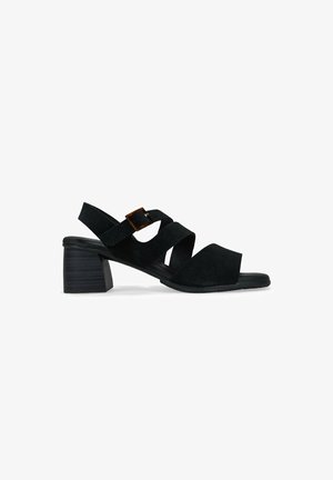 Sandalias de tacón de ante negro con correas cruzadas y un acento de hebilla. Tacón bajo y diseño de punta abierta. Textura suave, silueta minimalista.