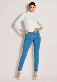 Frau in einem weißen grobstrickigen Rollkragenpullover, blauen hoch geschnittenen Skinny-Jeans, beigen High-Heels und goldenen Creolen, die mit Händen in den Hüften posiert.