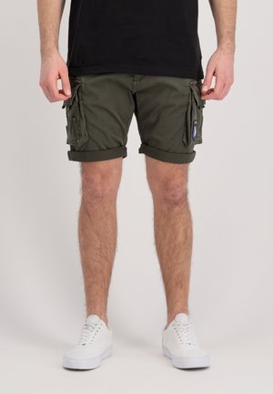 Homme portant un short cargo vert olive avec des ourlets retroussés, des baskets blanches et un t-shirt noir, debout devant un fond uni.