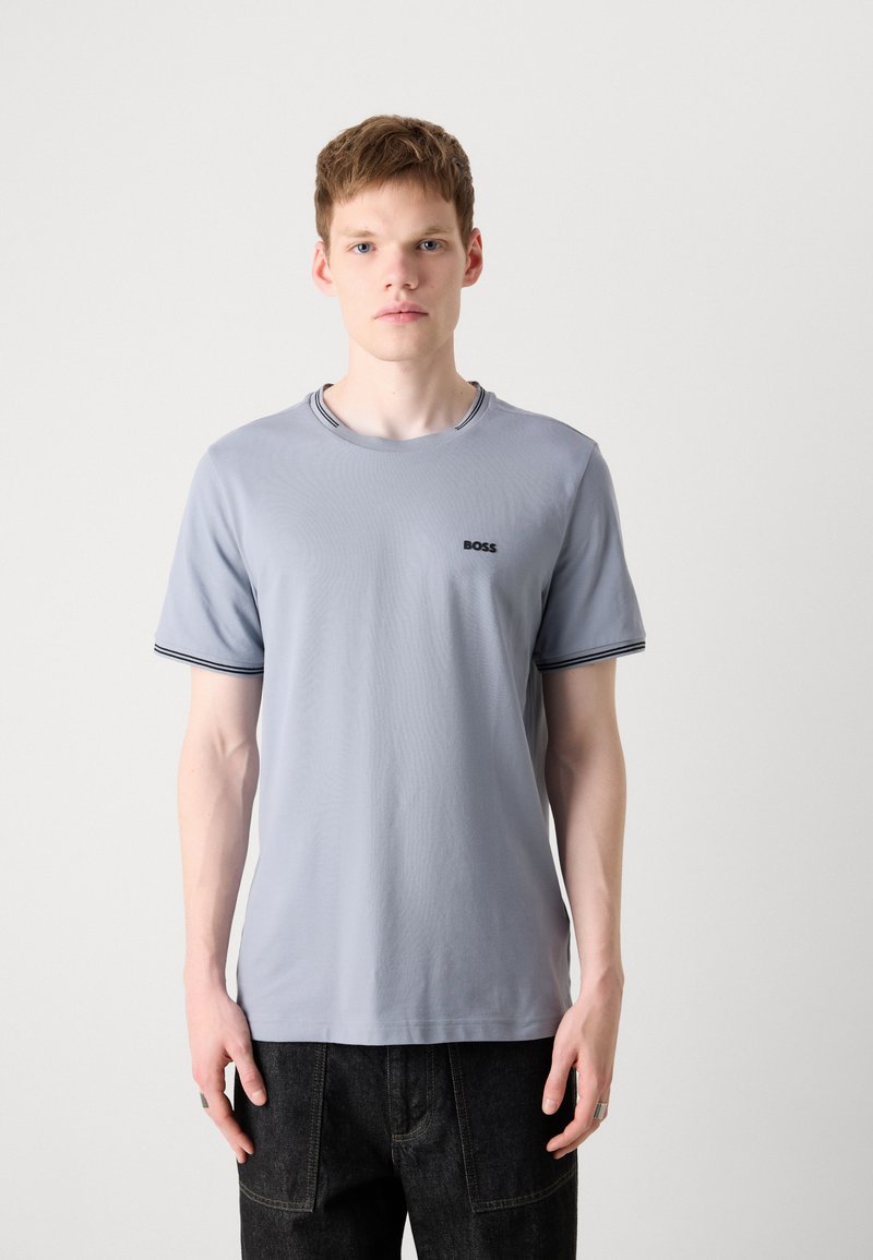 T-shirt gris à col rond avec logo noir, arborant une bordure rayée contrastante au niveau du col et des manches, fabriqué en tissu léger en mélange de coton.